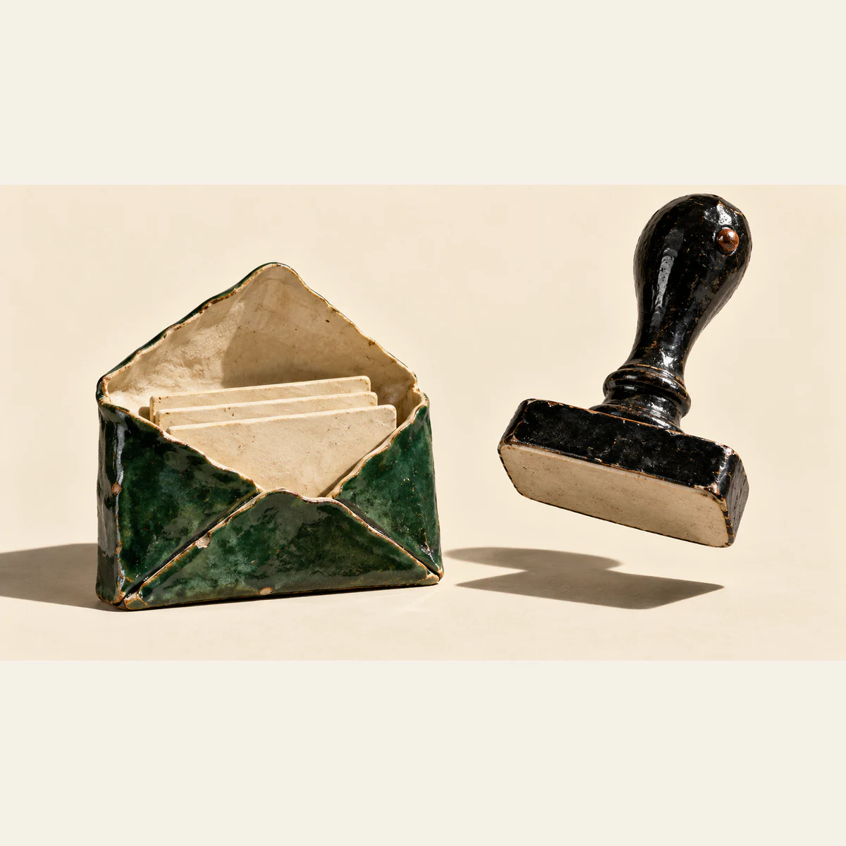 An open forest-green ceramic envelope holds blank ivory sheets beside a hovering black ceramic stamp with an unmarked underside. Chipped glaze and hard shadows punctuate the warm ivory background.