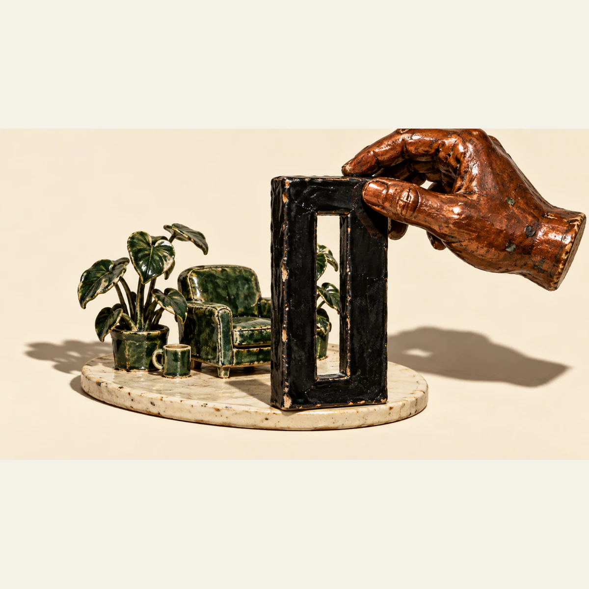 A burnt-copper ceramic hand holds a thick black frame with a narrow opening before a miniature green armchair, cup and houseplants. Chipped glazes and hard shadows texture the ivory scene.