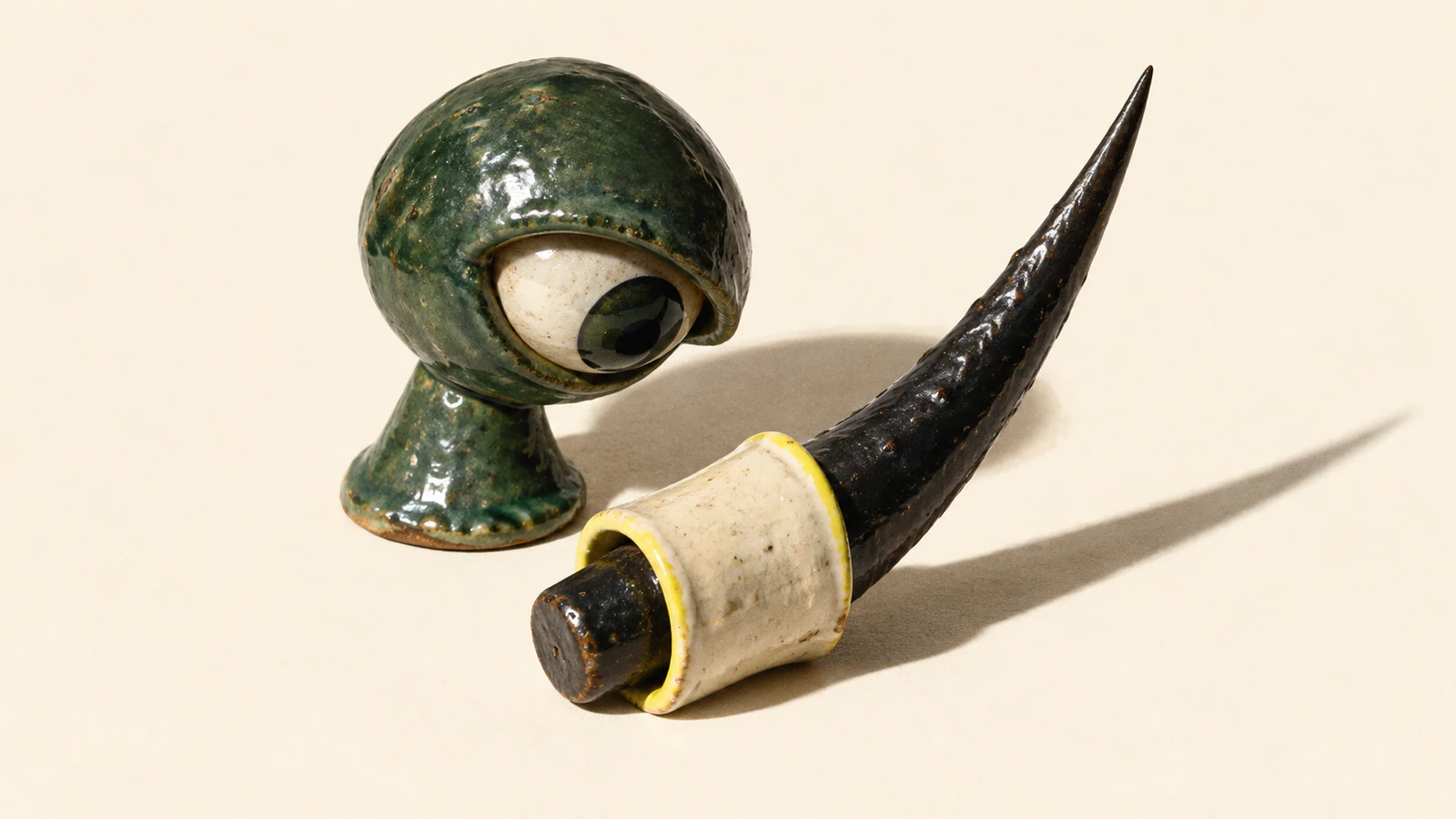 A mottled forest-green ceramic eye looks toward a yellow-edged ivory sleeve around a black ceramic thorn. Its long sharp tip remains exposed, casting a hard shadow across the warm ivory background.