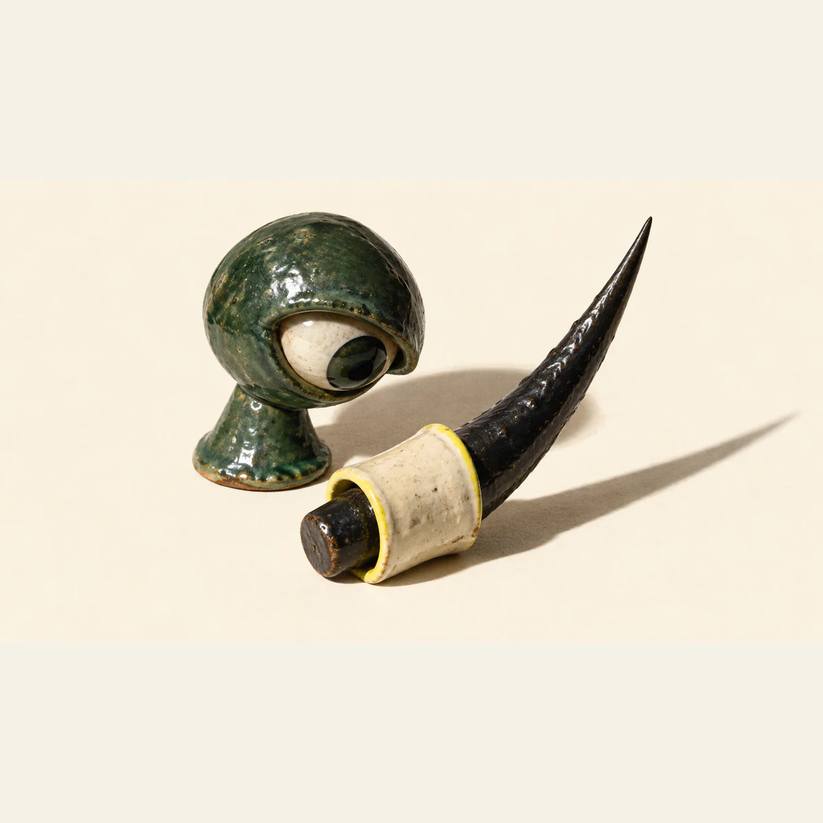 A mottled forest-green ceramic eye looks toward a yellow-edged ivory sleeve around a black ceramic thorn. Its long sharp tip remains exposed, casting a hard shadow across the warm ivory background.