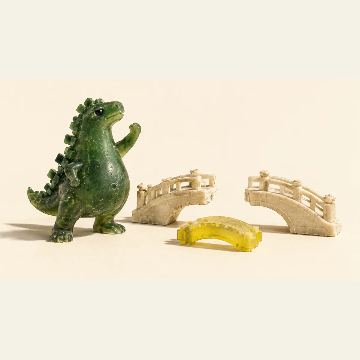 A textured green resin dinosaur raises one fist beside an ivory miniature bridge missing its middle span. A loose yellow bridge segment rests in front on a warm ivory background.