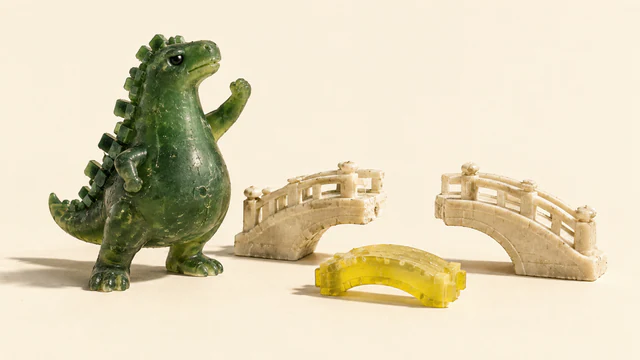 A textured green resin dinosaur raises one fist beside an ivory miniature bridge missing its middle span. A loose yellow bridge segment rests in front on a warm ivory background.