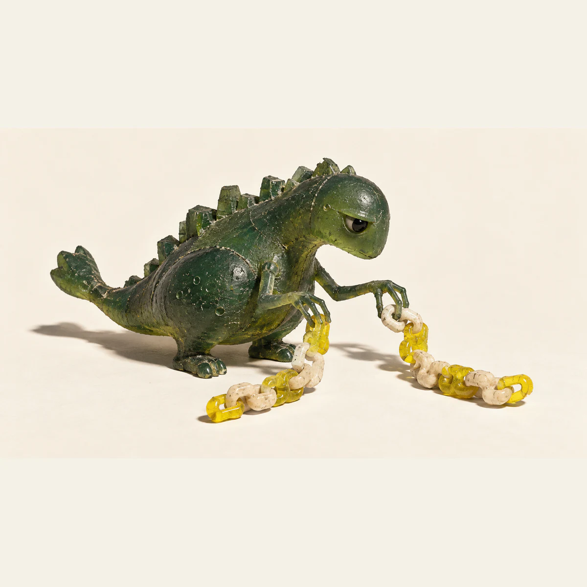 A skeptical green resin dinosaur holds two curling chains of ivory and yellow links arranged differently, casting hard shadows on a warm ivory background.