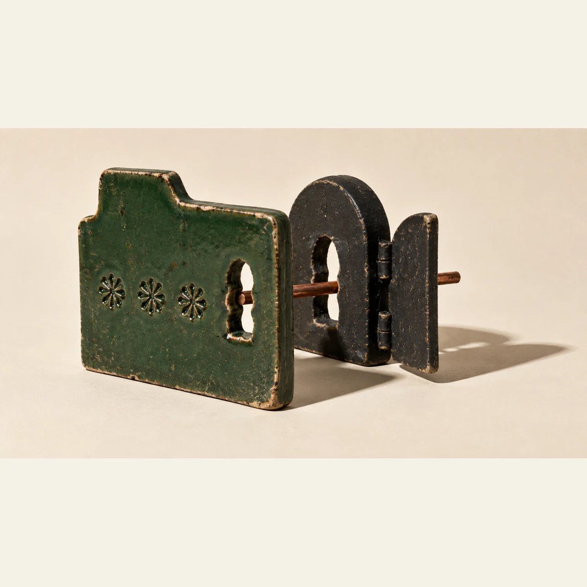 A copper-colored rod passes through aligned openings in a weathered green ceramic folder and a separate black ceramic doorway with an open shutter. Three flower impressions decorate the folder; hard shadows fall across an ivory background.